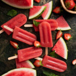 Assorted watermelon and strawberry popsicles are arranged on a dark surface, surrounded by slices of watermelon, halved strawberries, and mint leaves. This easy recipe is perfect for anyone looking to try delicious watermelon popsicles ideal for summer days.