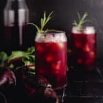 Two glass jars filled with iced, dark red beet lemonade and garnished with rosemary sprigs. A bottle and beetroot are in the background, all set against a dark surface.