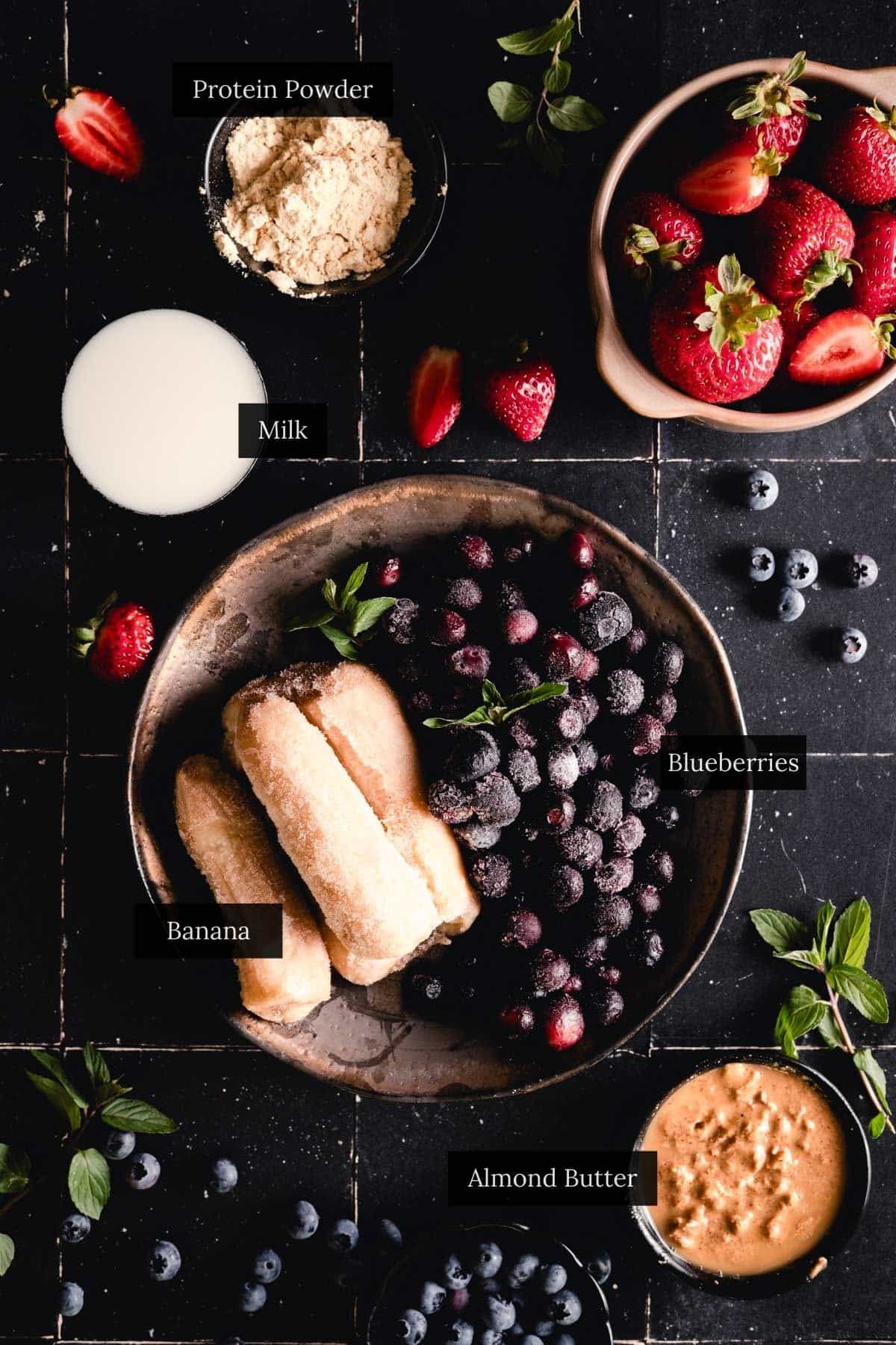Ingredients for homemade blueberry smoothie bowl on a black tiled counter.