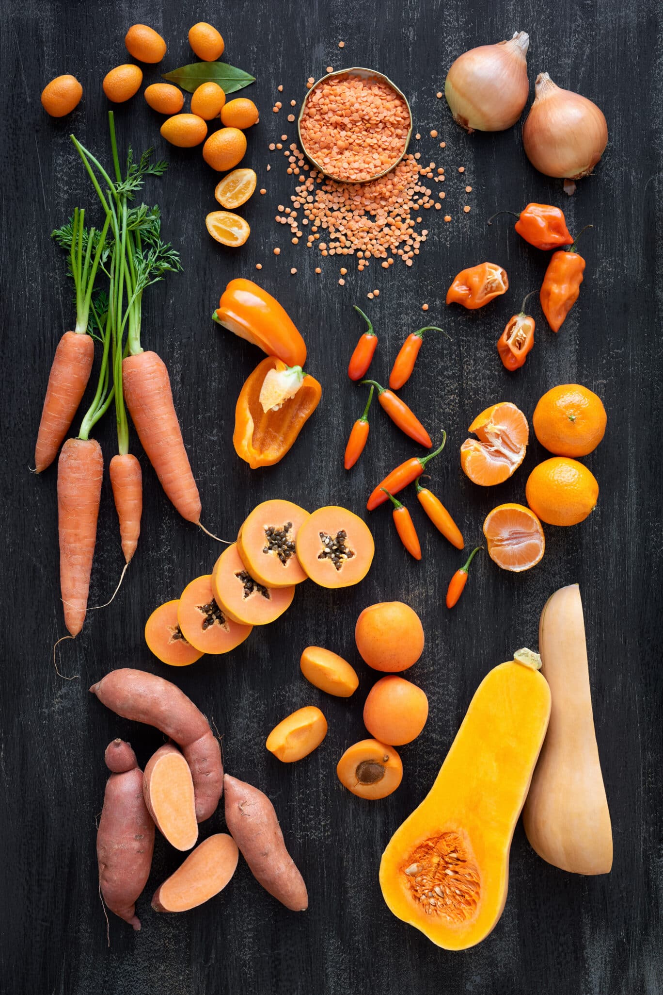 Orange foods and snacks on a black surface including red lentils, squash, sweet potatoes, carrots, and more.