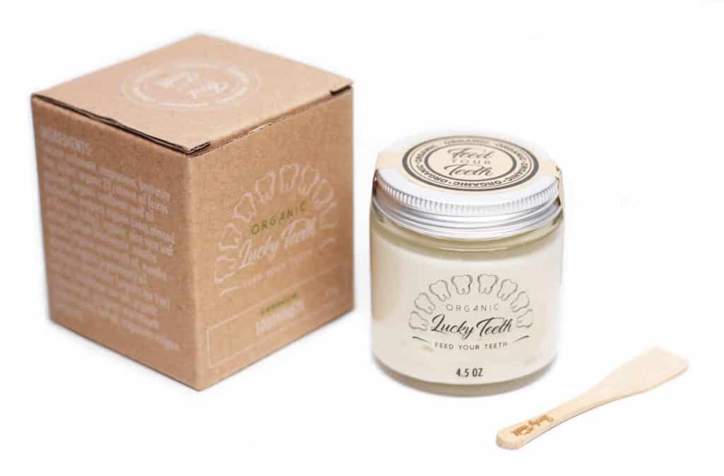 Glass jar with metal lid of toothpaste next to a bamboo scoop and a kraft box.