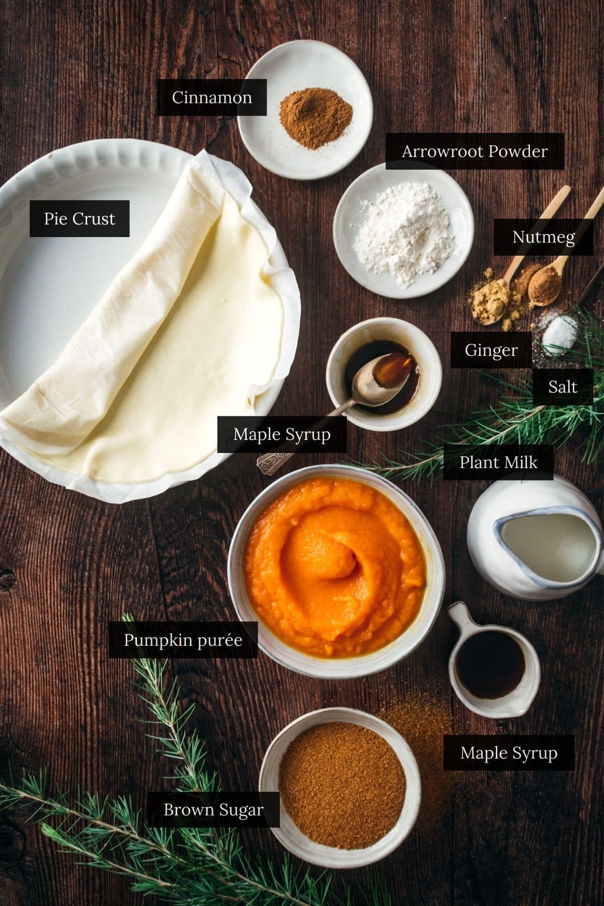 Ingredients for a delicious vegan pumpkin pie on a dark wooden surface, including pie crust, pumpkin purée, spices, plant milk, brown sugar, maple syrup, and arrowroot powder—all carefully labeled.