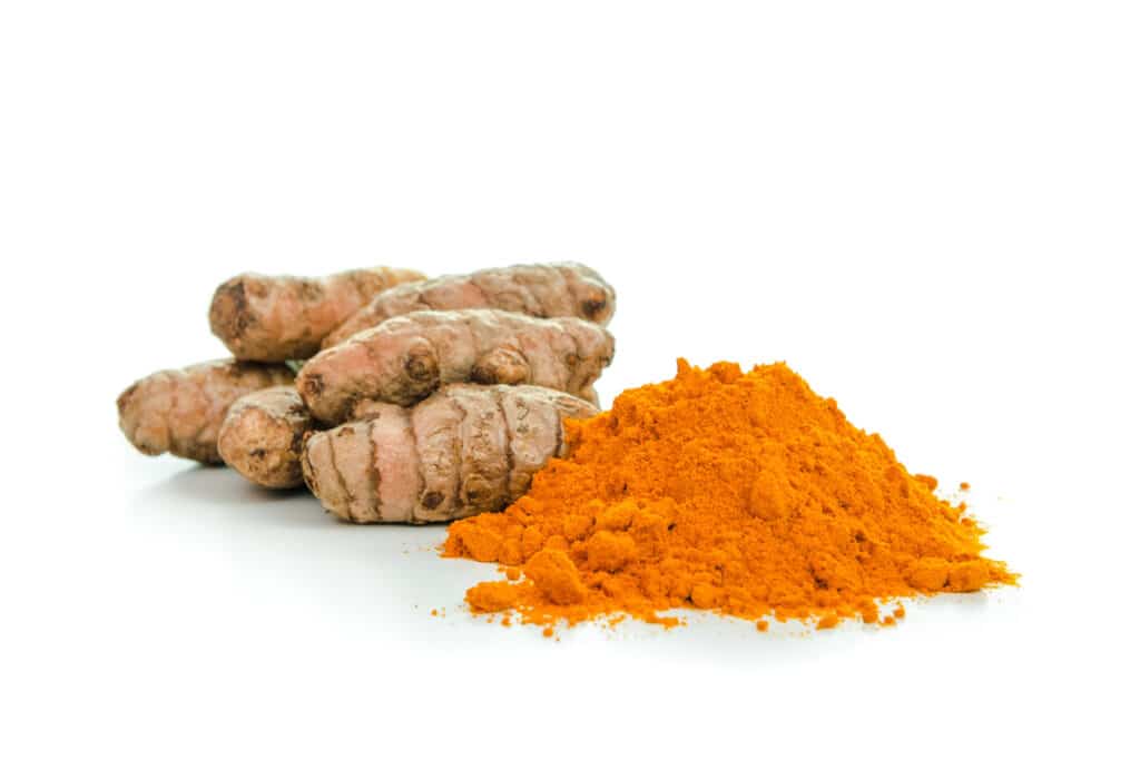 Fresh turmeric root with a pile of dried turmeric powder on a white surface.