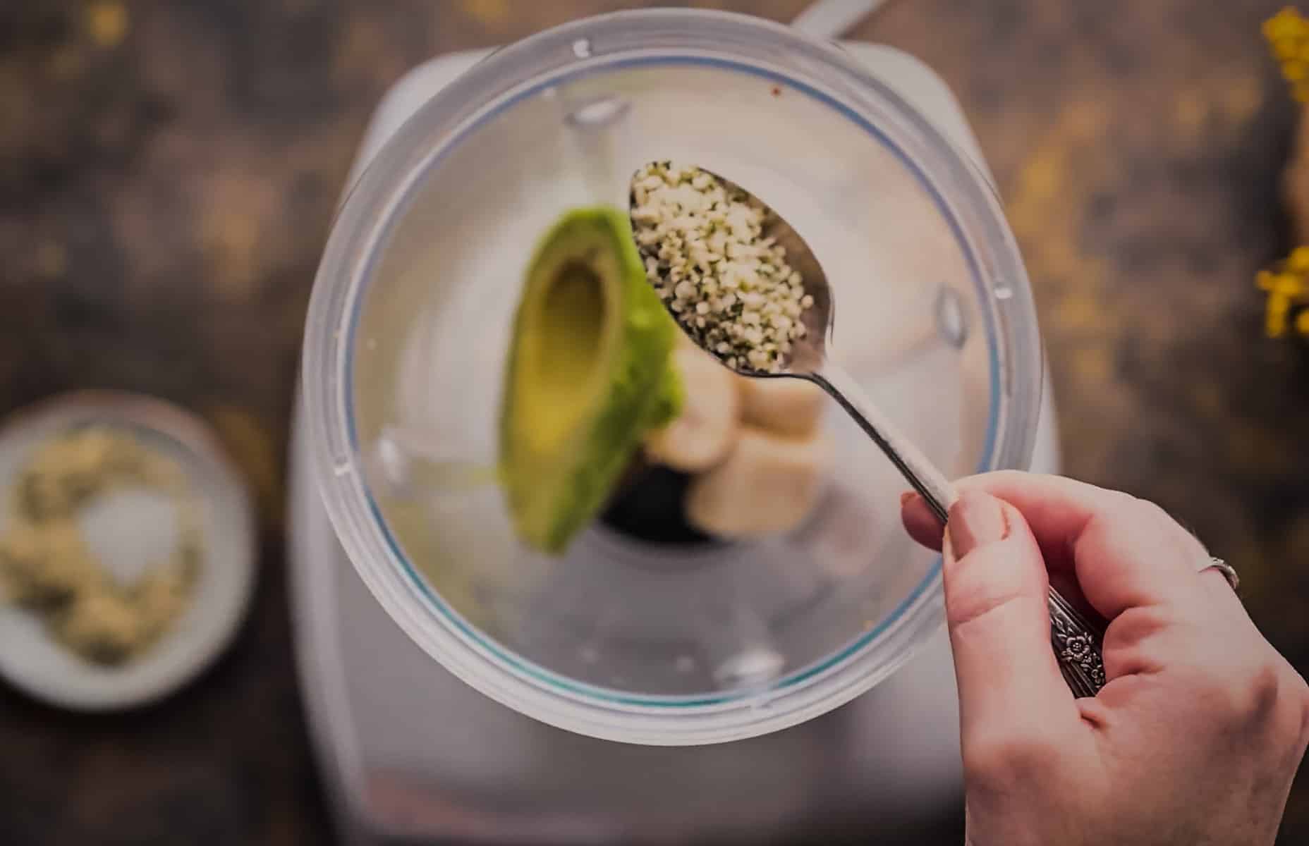 A hand holds a spoonful of hemp hearts above a blender.