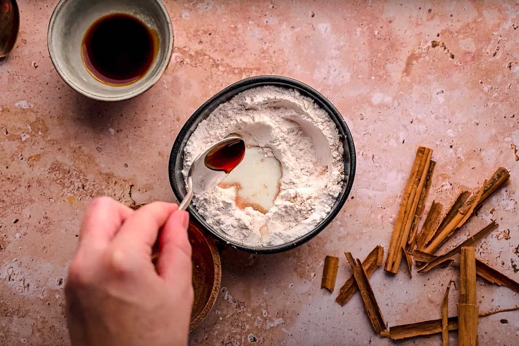 A hand holds a spoon of vanilla extract over a bowl of powdered sugar.