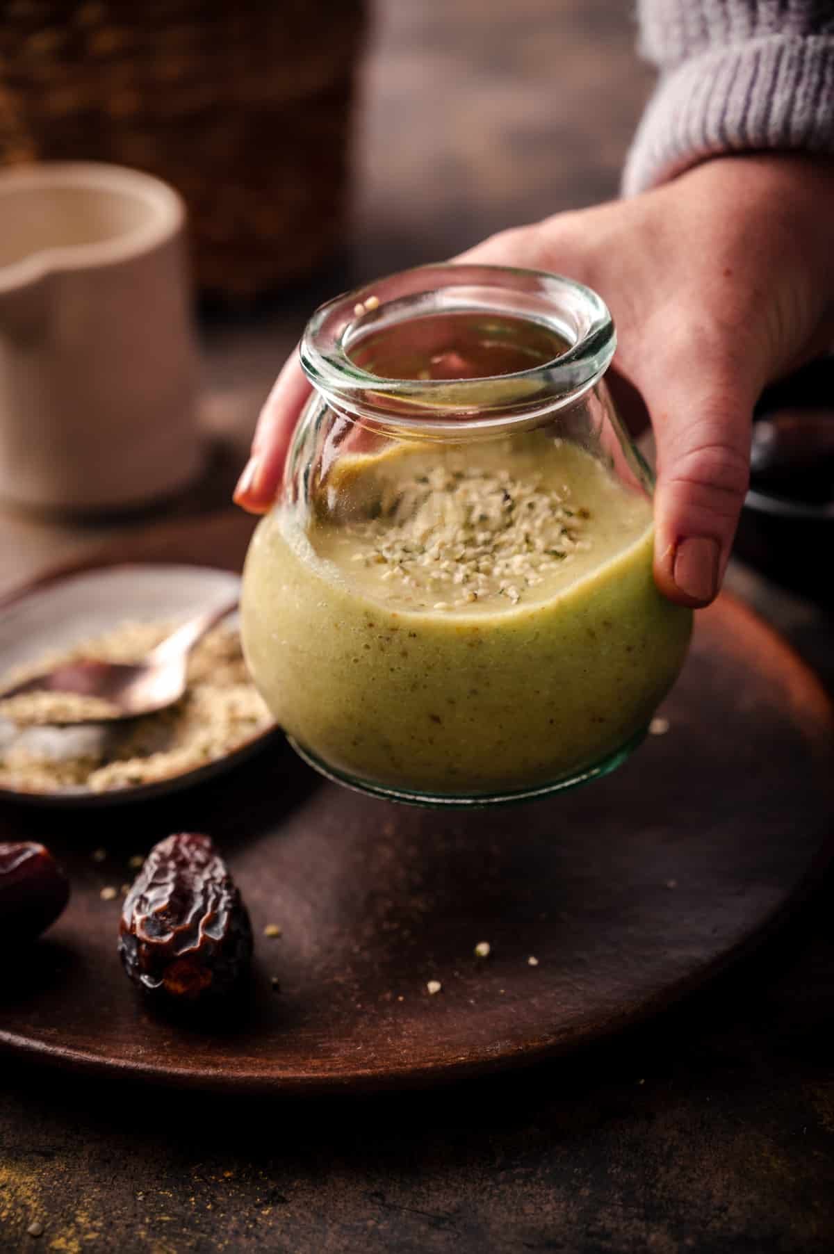 Avocado smoothie in a glass cup, hemp seeds sprinkled on top being picked up by a woman.