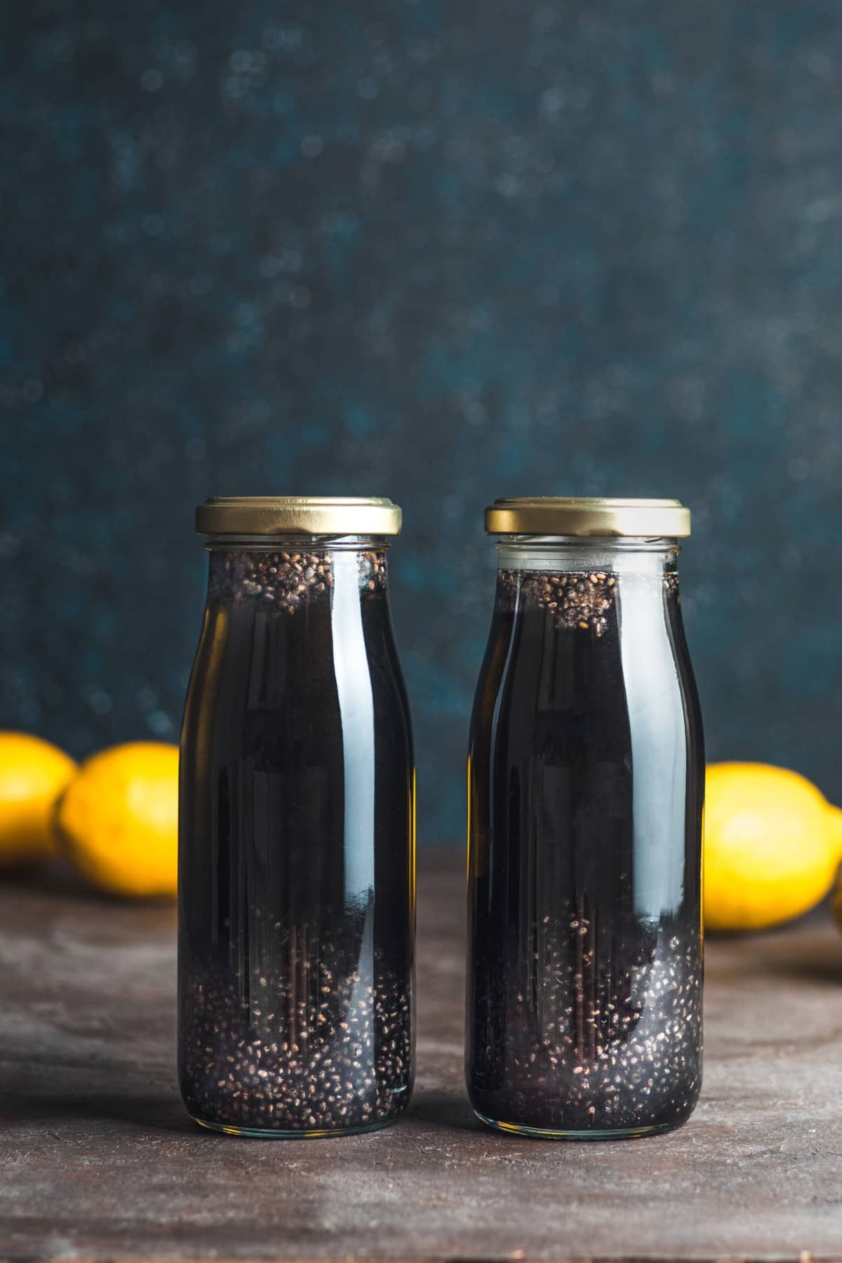 Two glass bottles of charcoal lemon chia drink with lemons behind it.