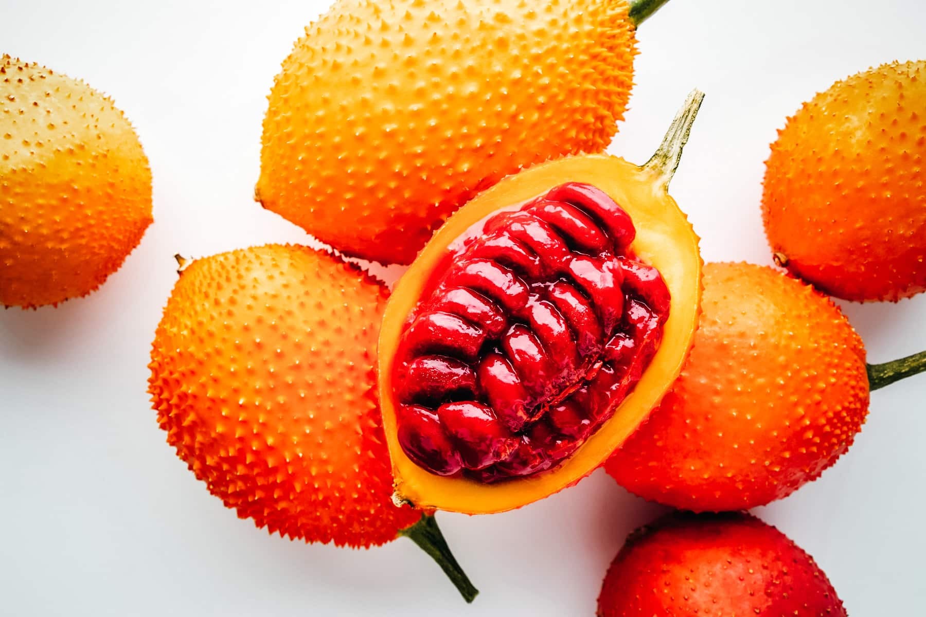 Several orange gac fruits with one cut in half showing its red colored seeds.