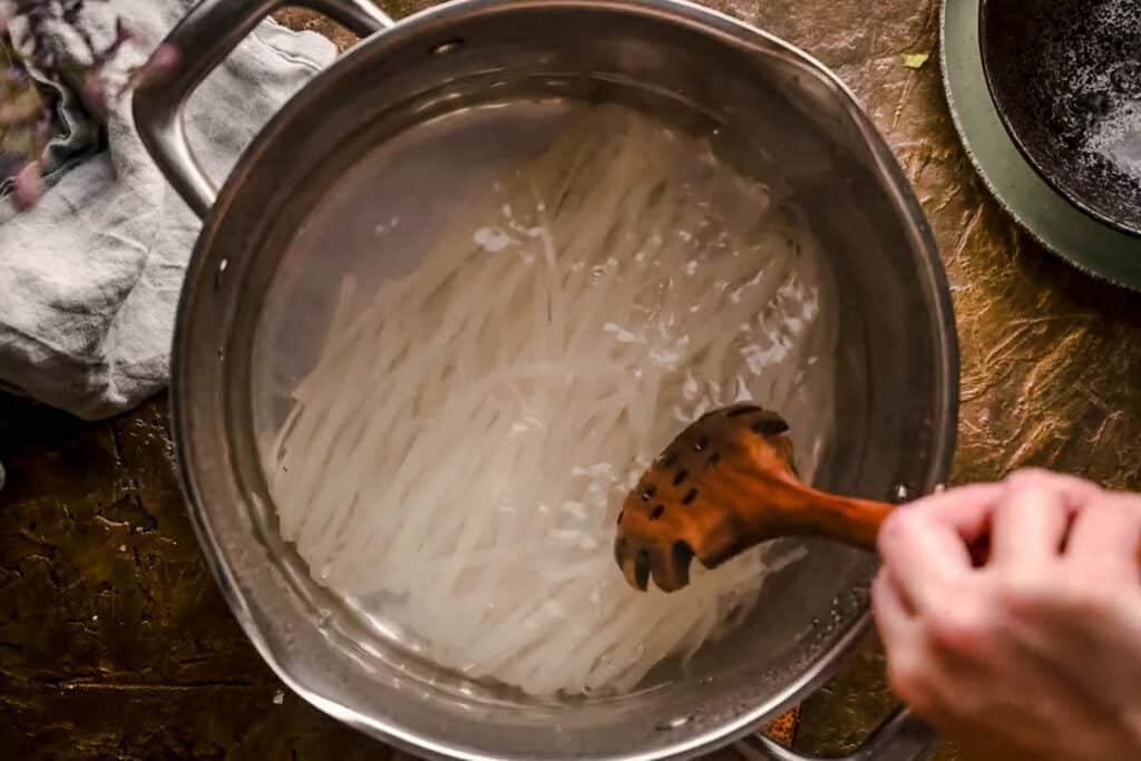 Wooden spoon stirring pot of rice noodles.