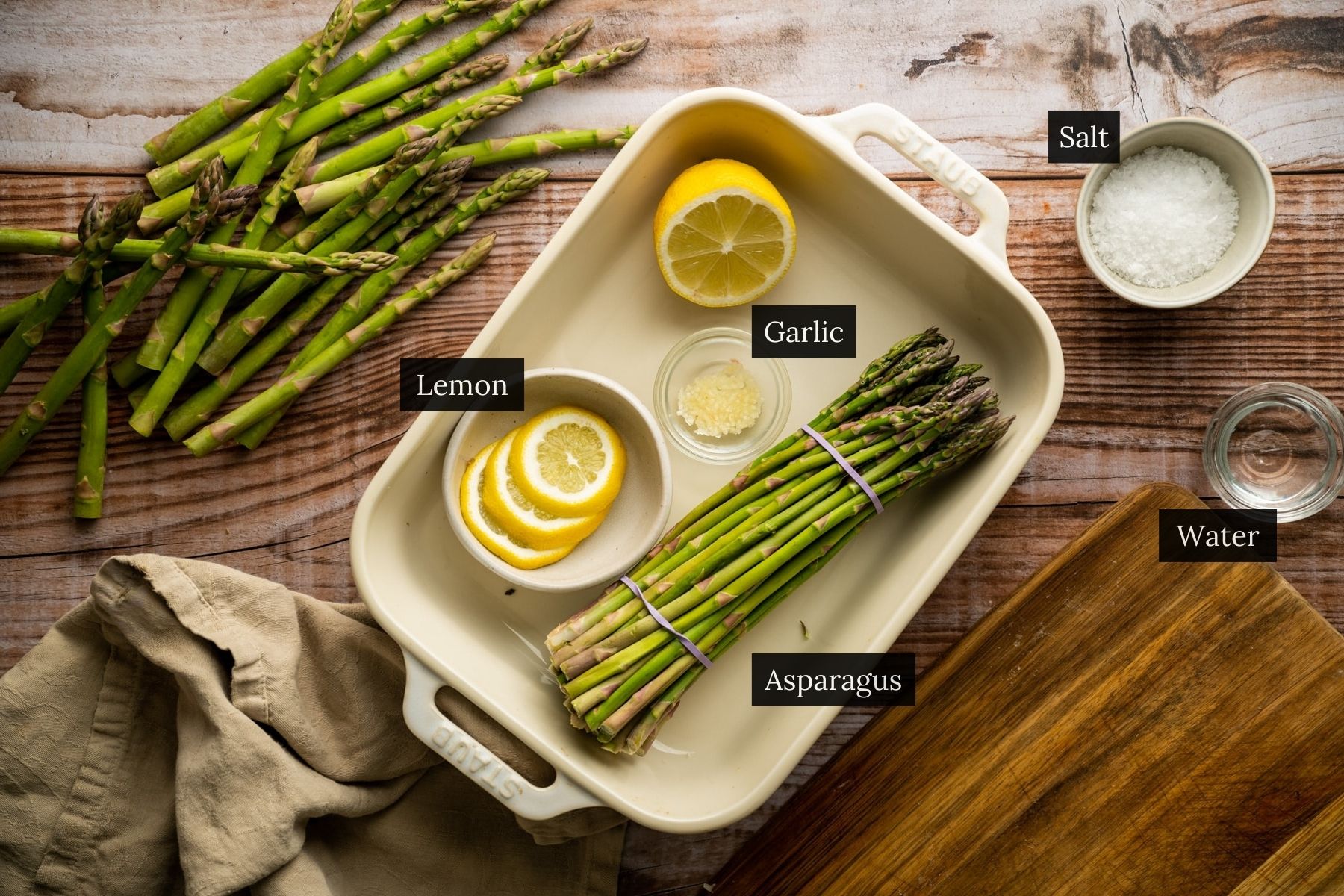 Ingredients for roasting asparagus are arranged on a wooden surface, including asparagus, lemon, garlic, salt, and water. For those wondering how to microwave asparagus instead, you can substitute the wooden board for a microwave-safe dish and maintain similar ingredients.