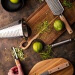 A tabletop displays a variety of kitchen tools including graters, a cutting board, a knife, and a peeler, alongside whole and grated limes with zest scattered around.