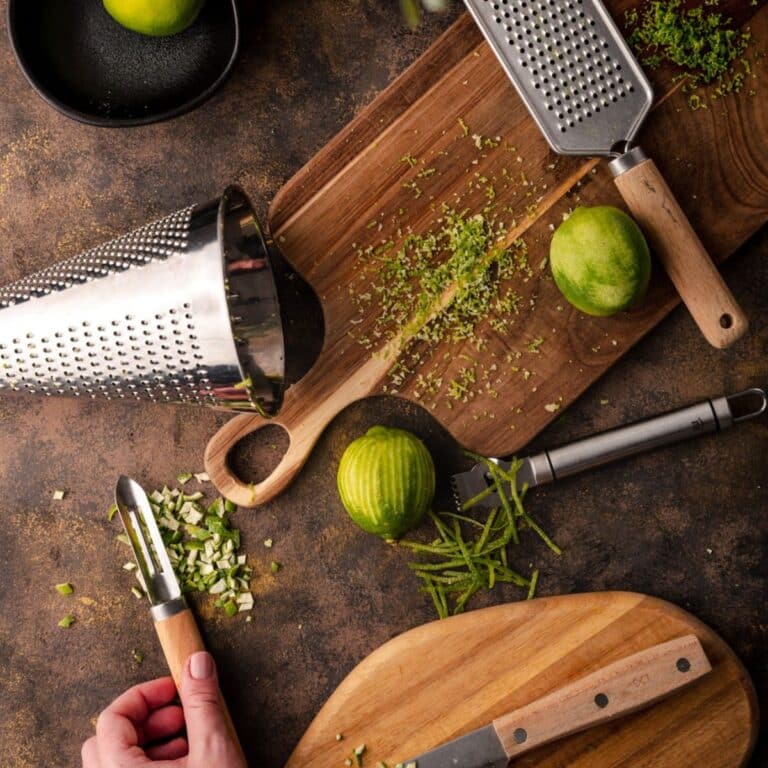A tabletop displays a variety of kitchen tools including graters, a cutting board, a knife, and a peeler, alongside whole and grated limes with zest scattered around.