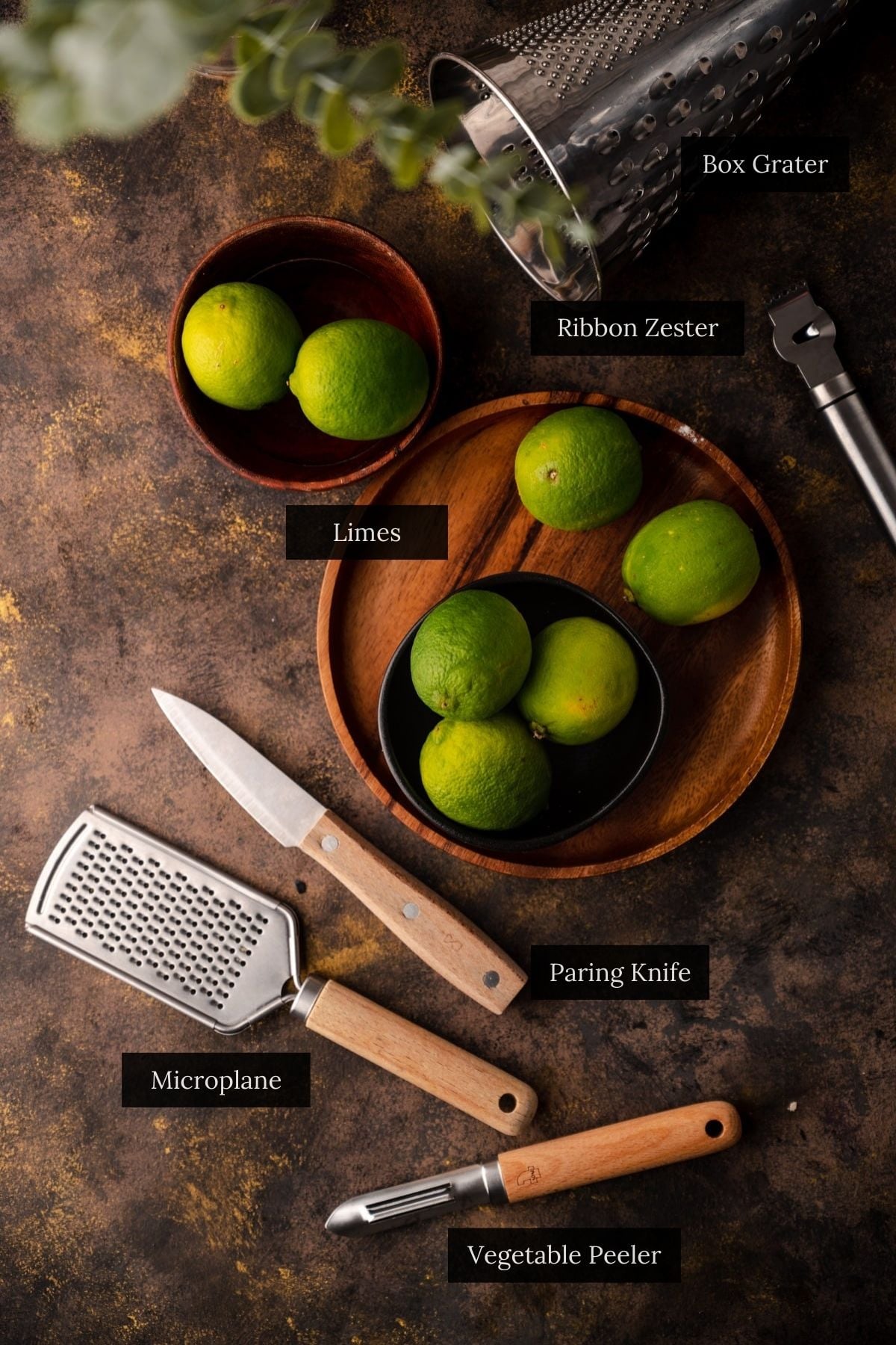 An assortment of kitchen tools including a box grater, ribbon zester, microplane, paring knife, and vegetable peeler, and limes.