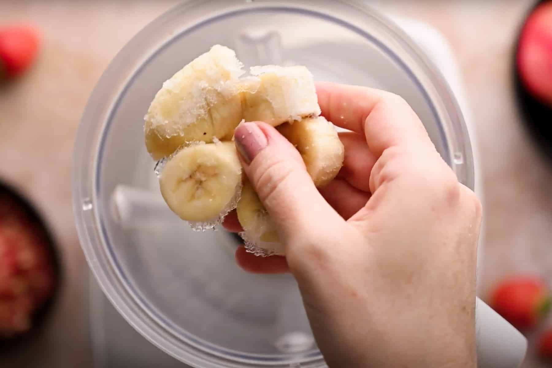 A hand holding frozen banana slices over a blender container, preparing to make a pomegranate smoothie. 