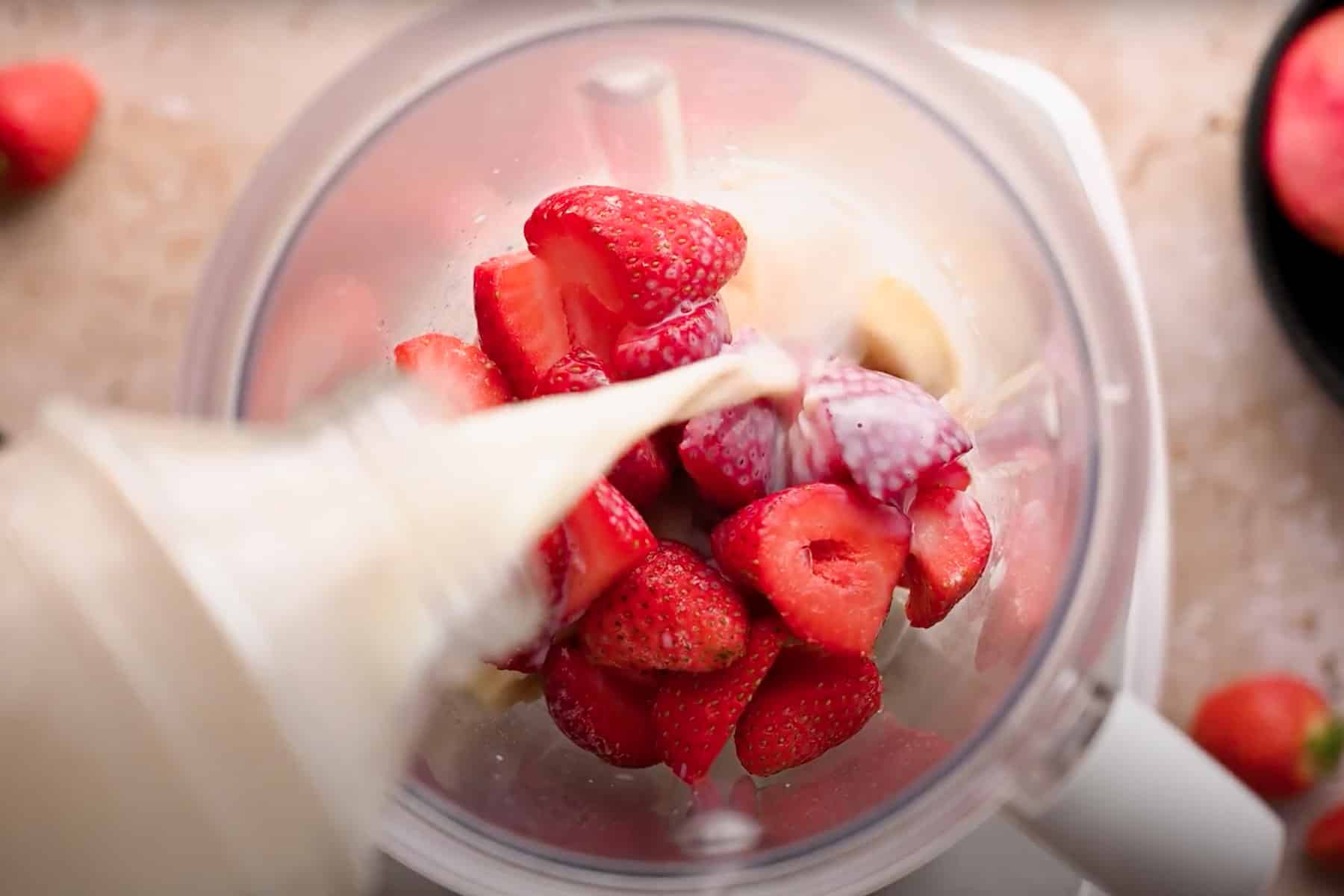 A blender being filled with fresh strawberries and oat milk, being poured in.