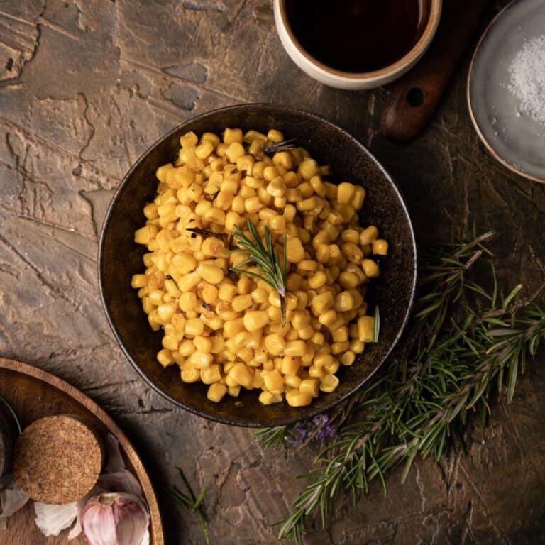 A bowl of cooked corn, garnished with a sprig of rosemary and placed on a bronze textured surface.