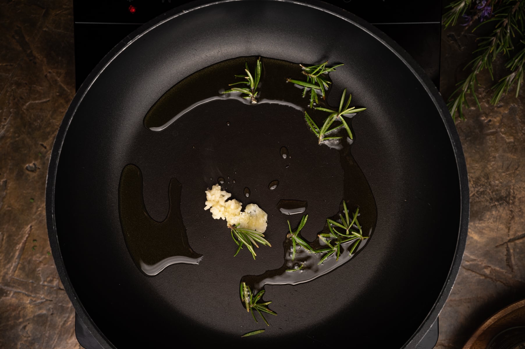 A frying pan with olive oil, minced garlic, and rosemary sprigs.