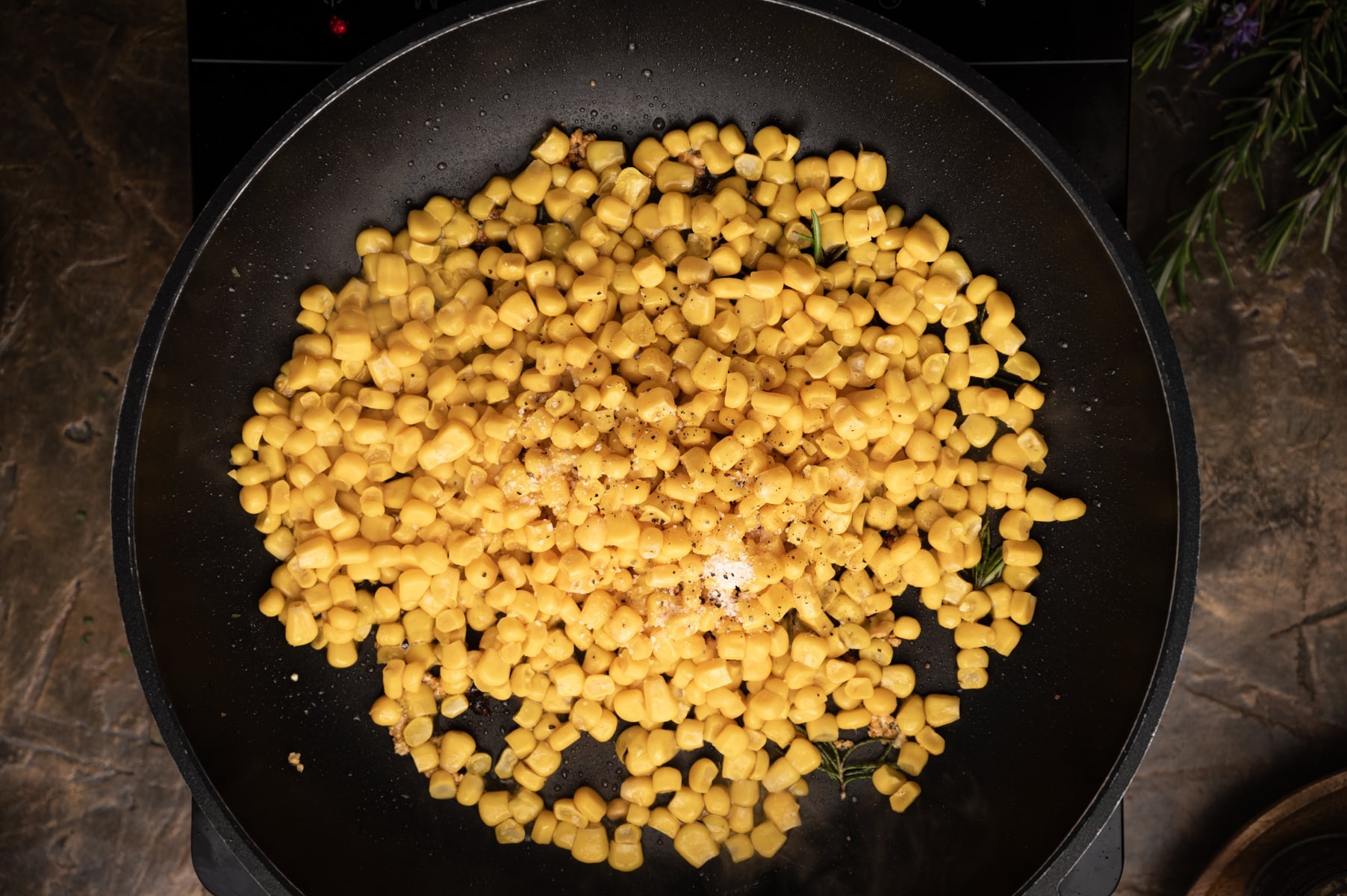 A black frying pan filled with cooked corn kernels, seasoned with spices, on a dark countertop.