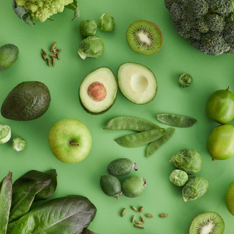 An assortment of fruits that are green, including avocado, kiwi, lime, apple, lettuce, and snap peas, elegantly arranged on a vibrant green background.