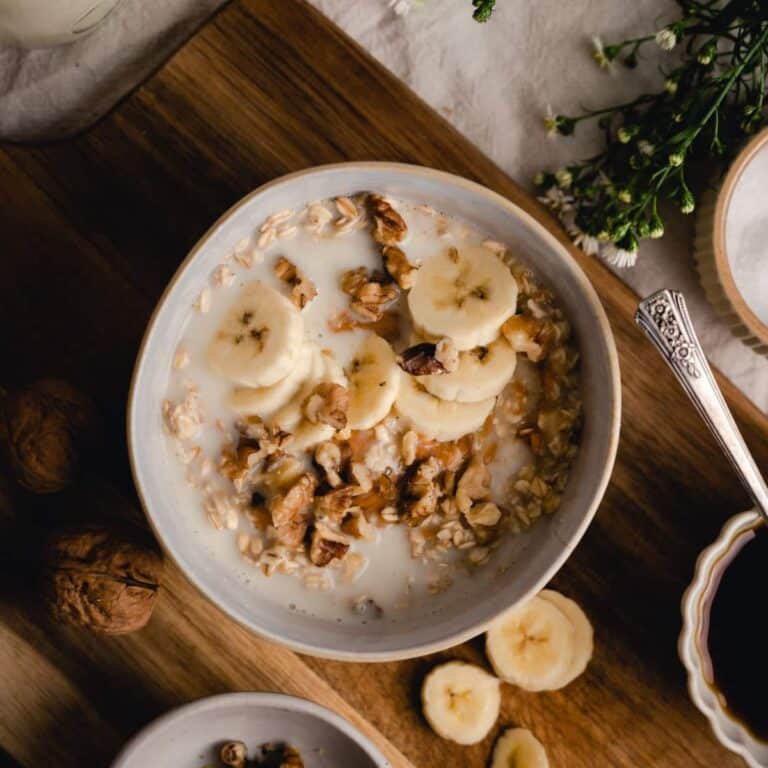 A bowl of banana oatmeal topped with nuts, set on a wooden board with a spoon.