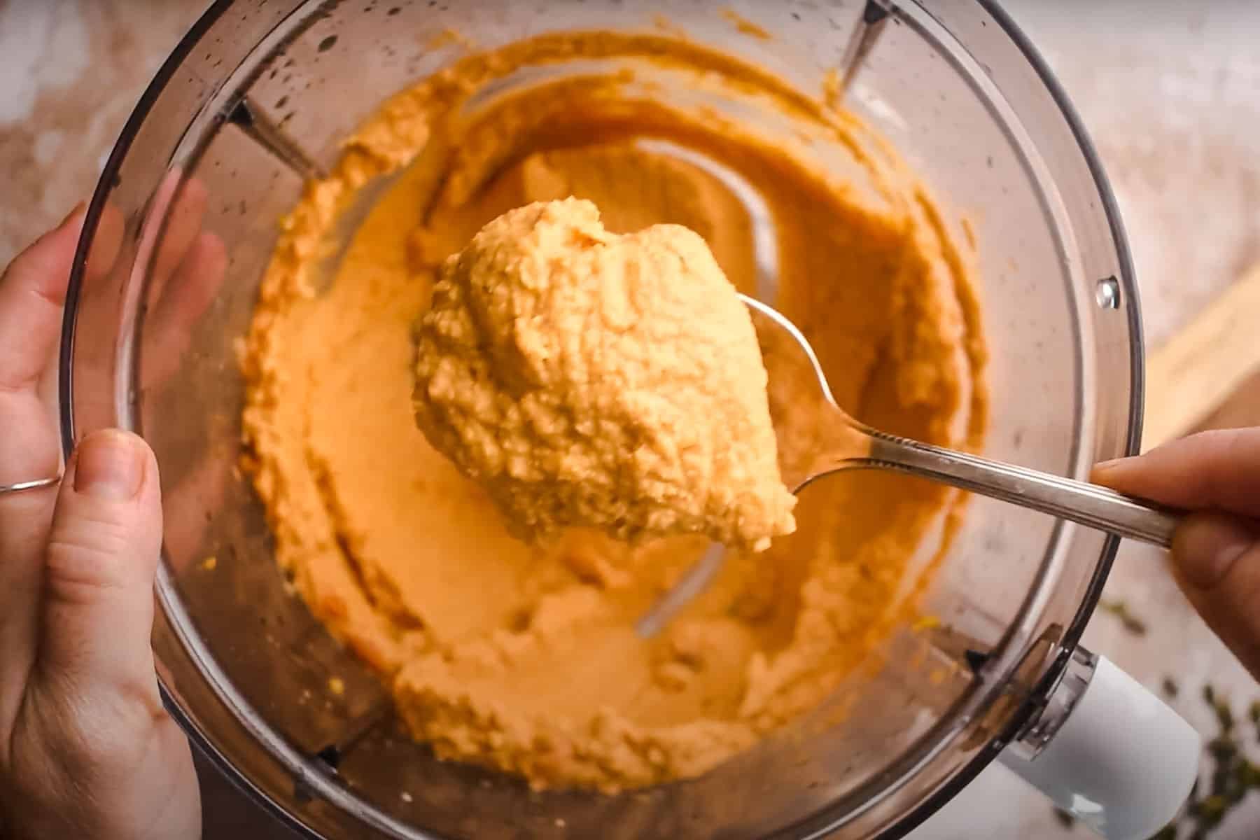 A person holding a spoonful of blended pumpkin hummus above a food processor bowl.