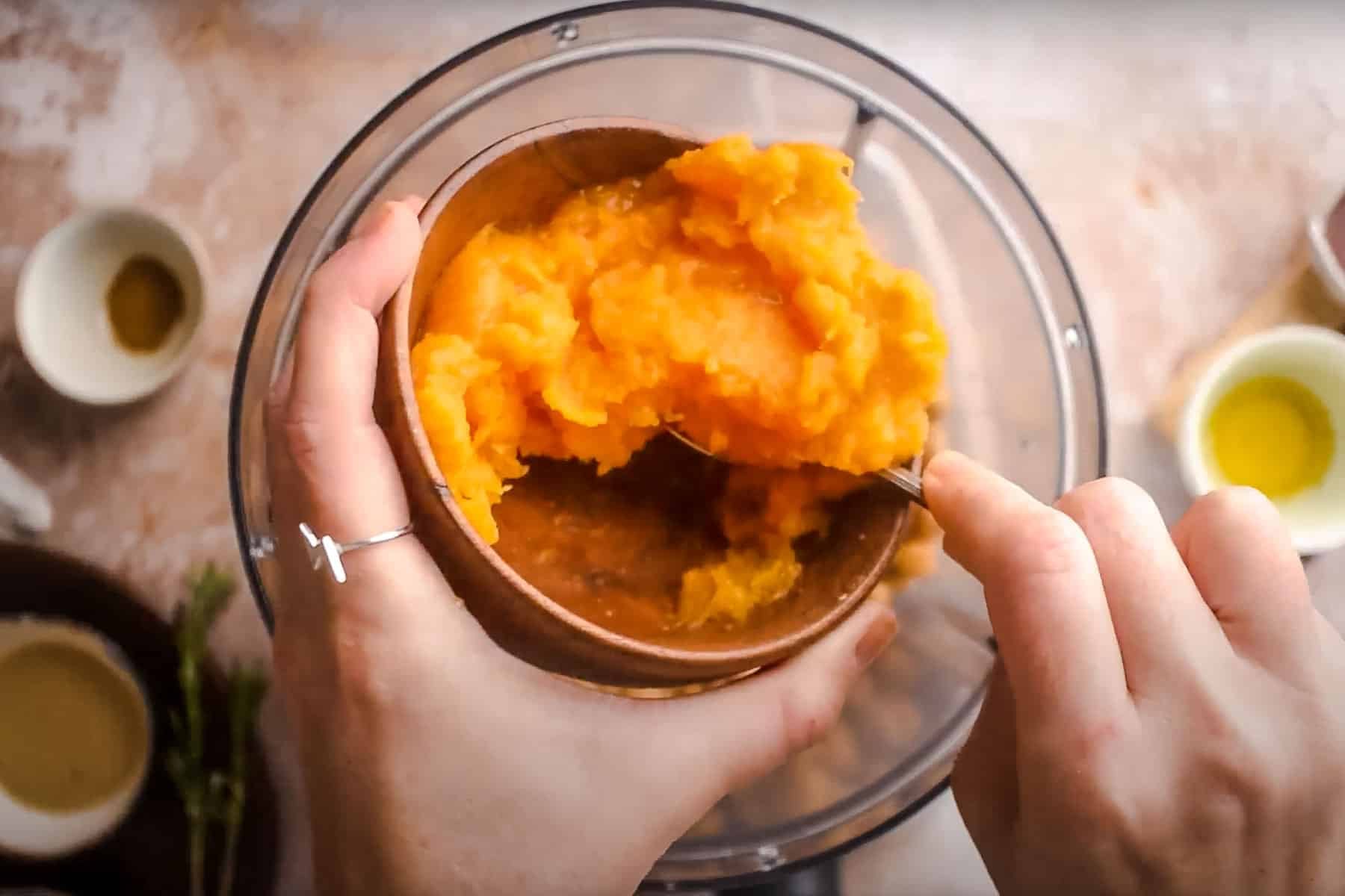 Hands holding a brown bowl filled with pumpkin puree, preparing to transfer them into a food processor.