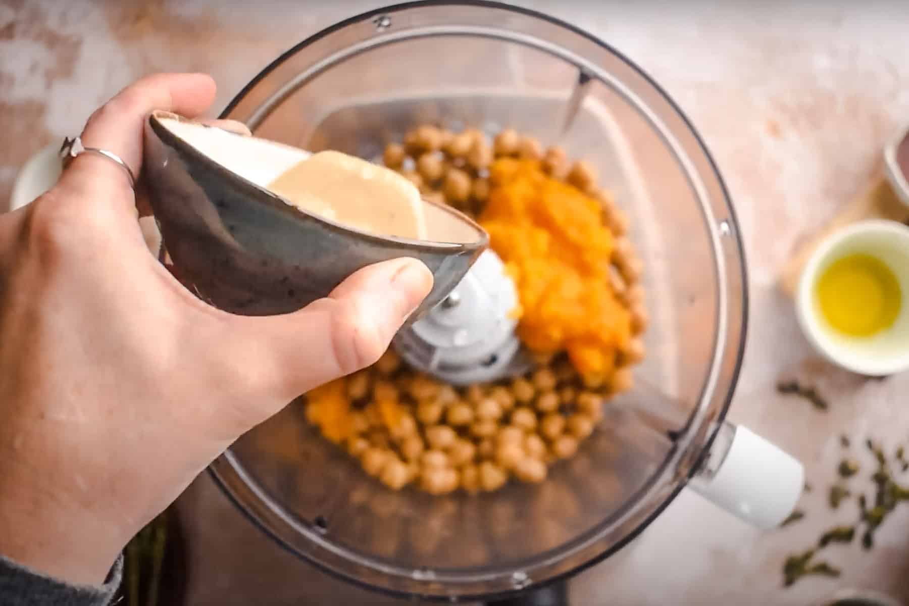 A person pours tahini into a food processor containing chickpeas and pumpkin puree.