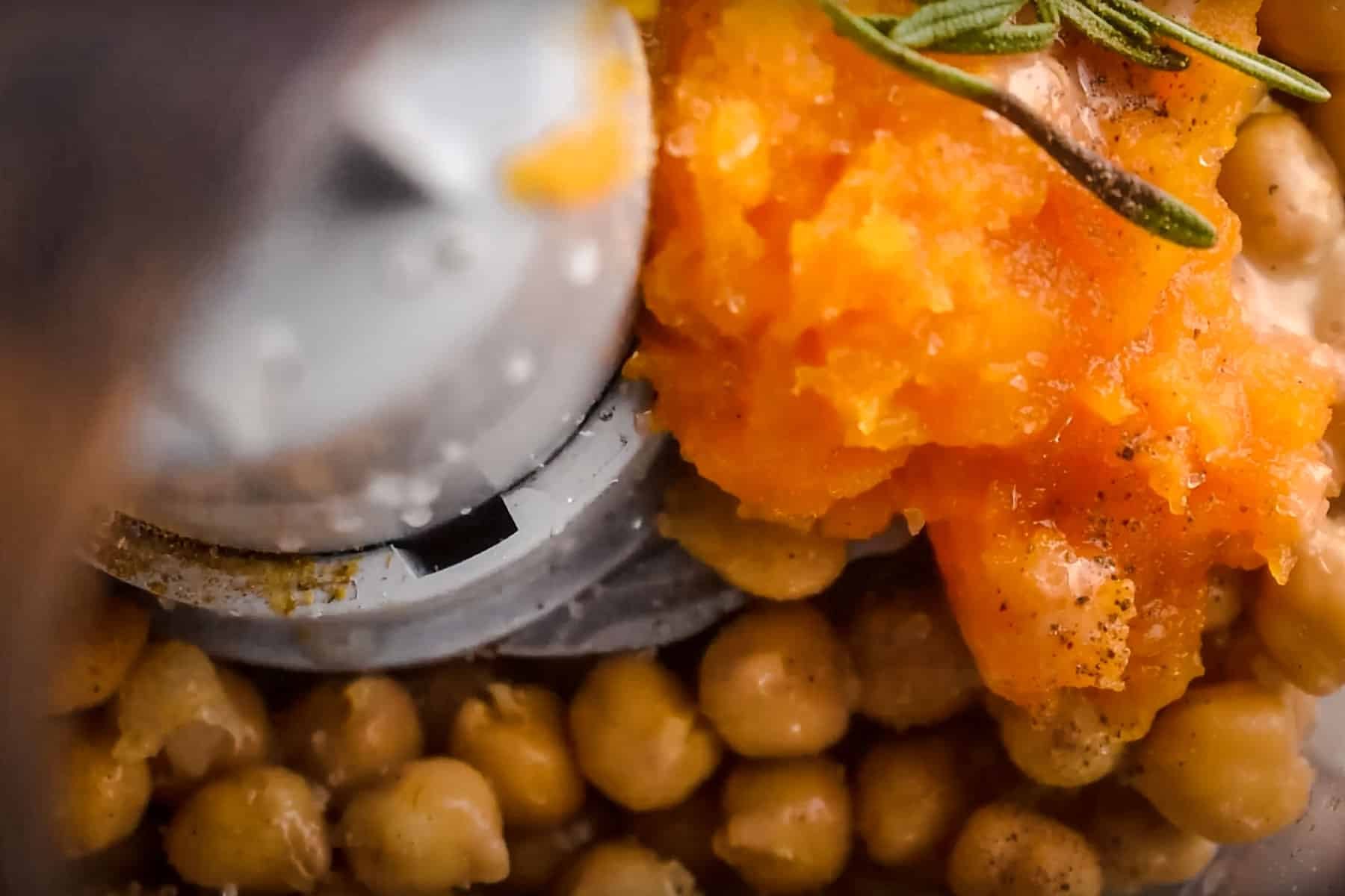 Close-up of a food processor containing chickpeas, a dollop of tahini, and a sprig of rosemary, and more.