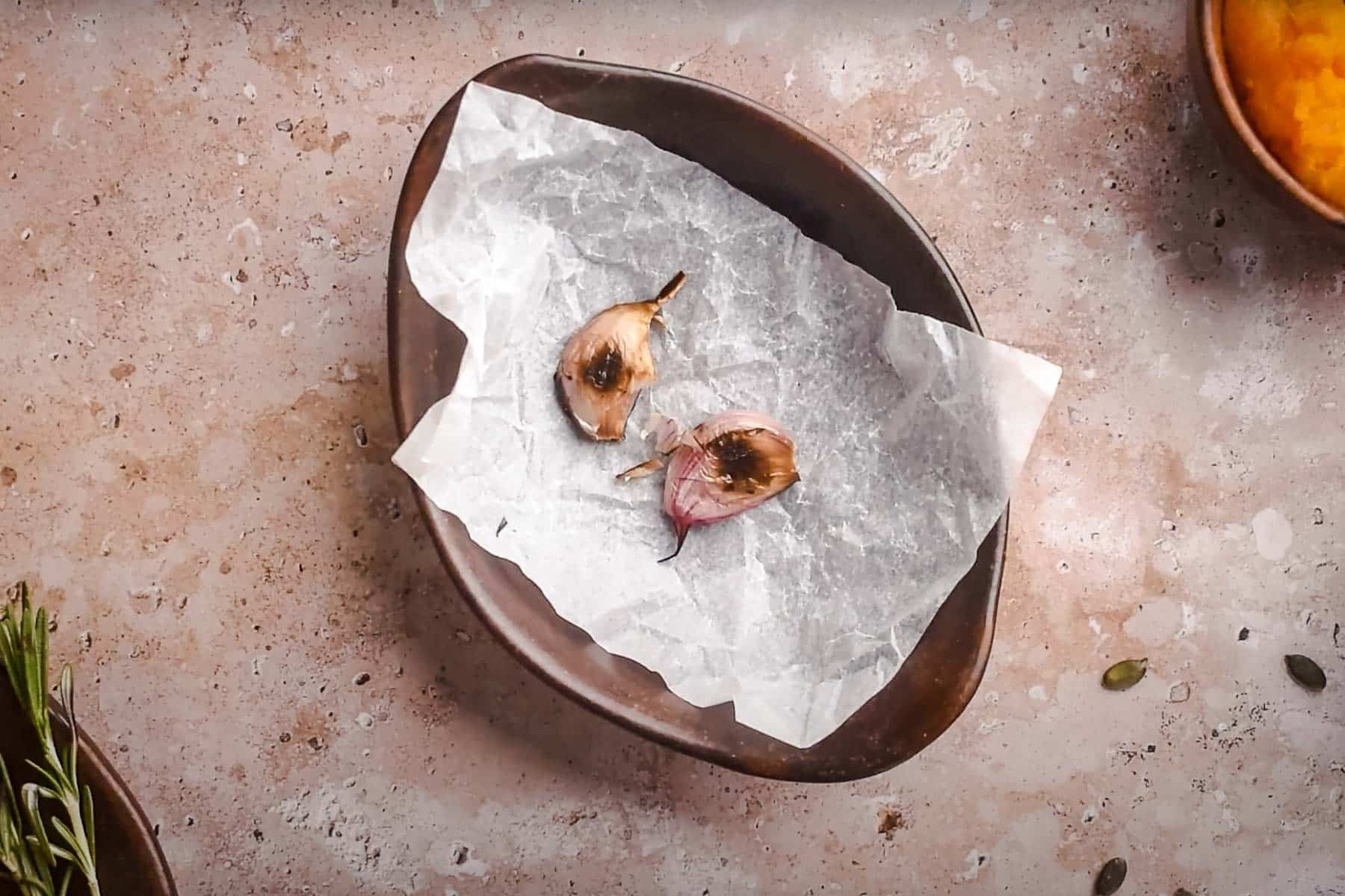 Two roasted garlic cloves rest on a piece of parchment paper in a dark brown bowl.
