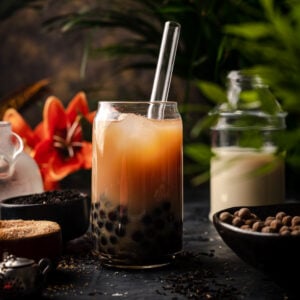 A glass of bubble tea with tapioca pearls and ice, served with a clear straw, surrounded by tea ingredients, sugar, a jar of milk, and a red flower in the background—nothing beats this classic milk tea recipe.