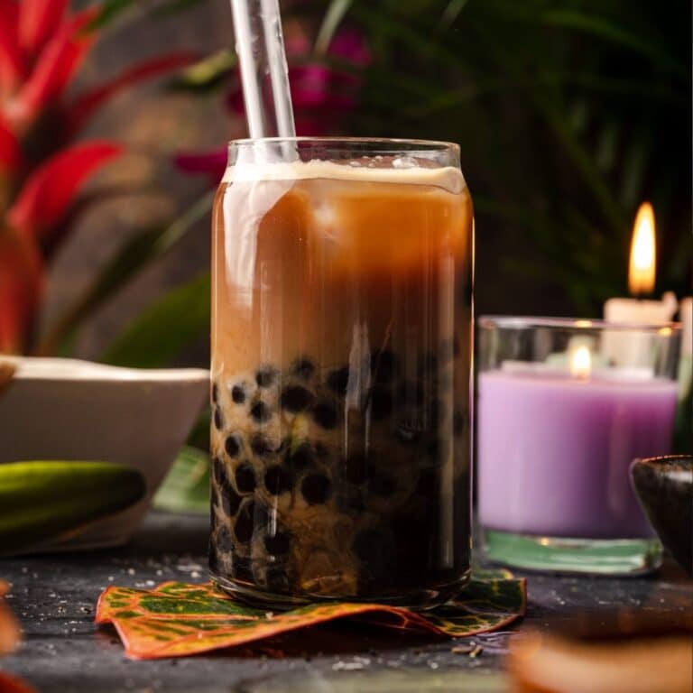 A glass of coffee boba with a straw, nestled on a leaf, surrounded by a lit candle and lush tropical plants.