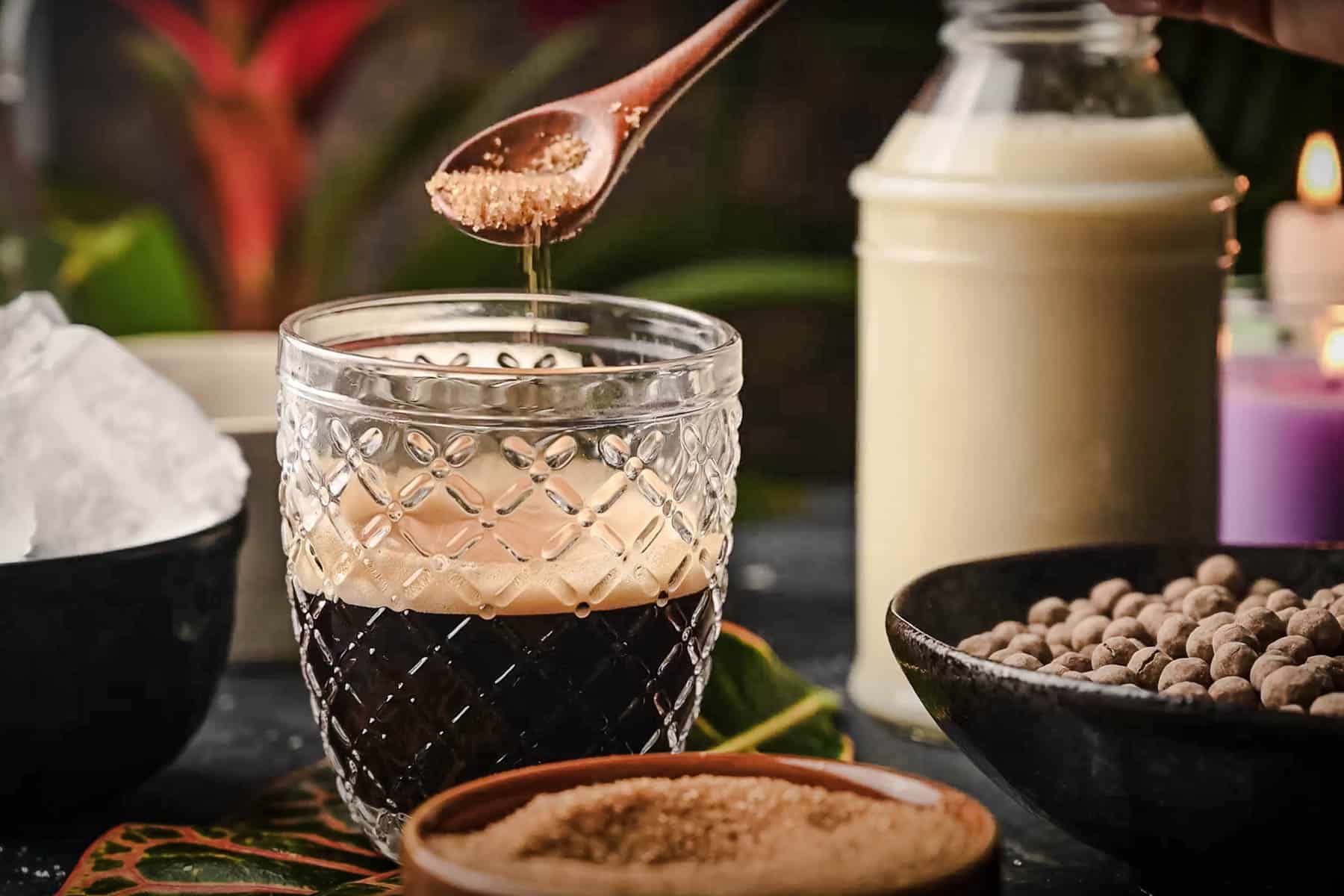 A spoon adds sugar to a glass of iced coffee boba next to a bottle of milk, candles, ice, and spices on the table.