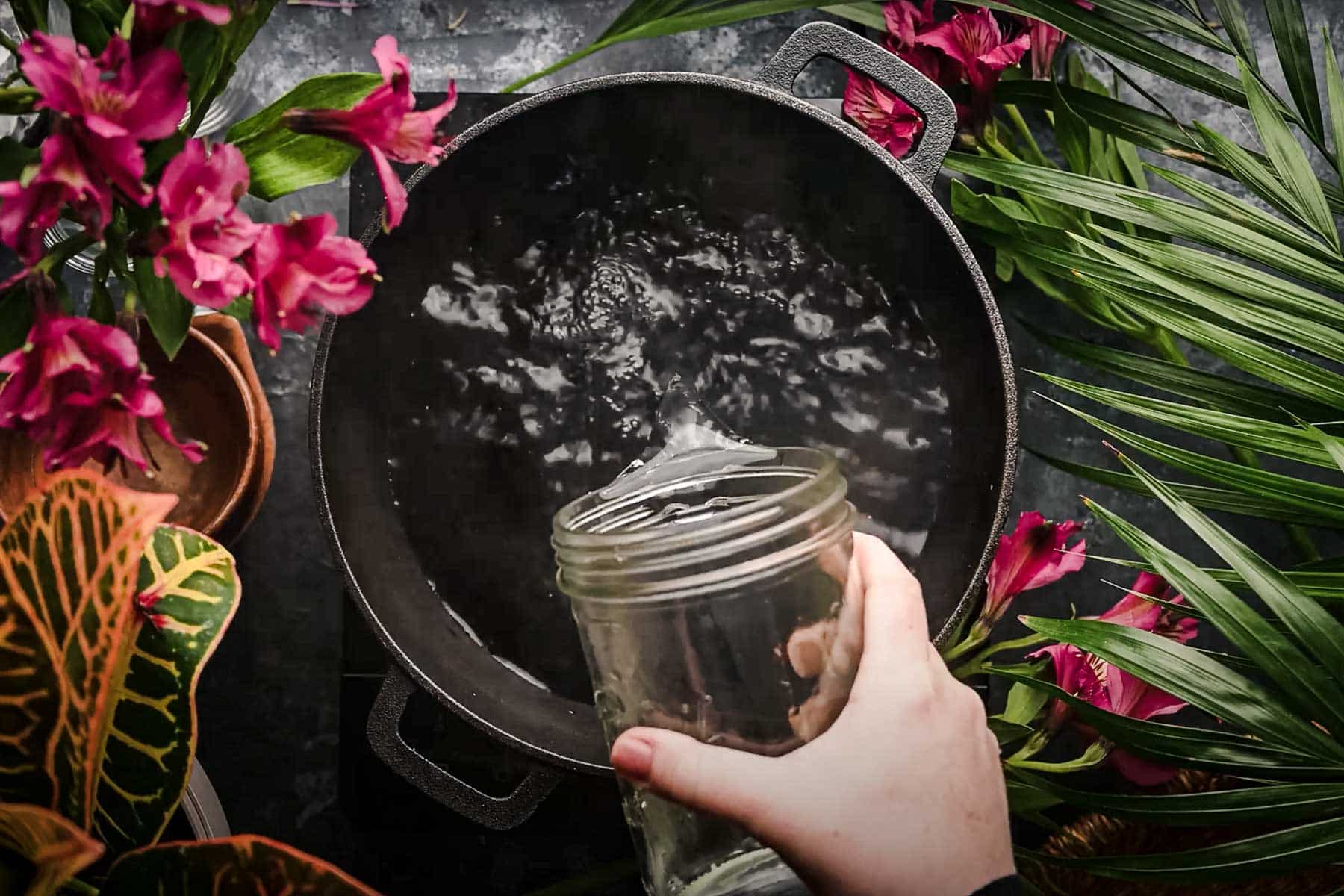 A hand pouring water from a glass jar into a black pot.