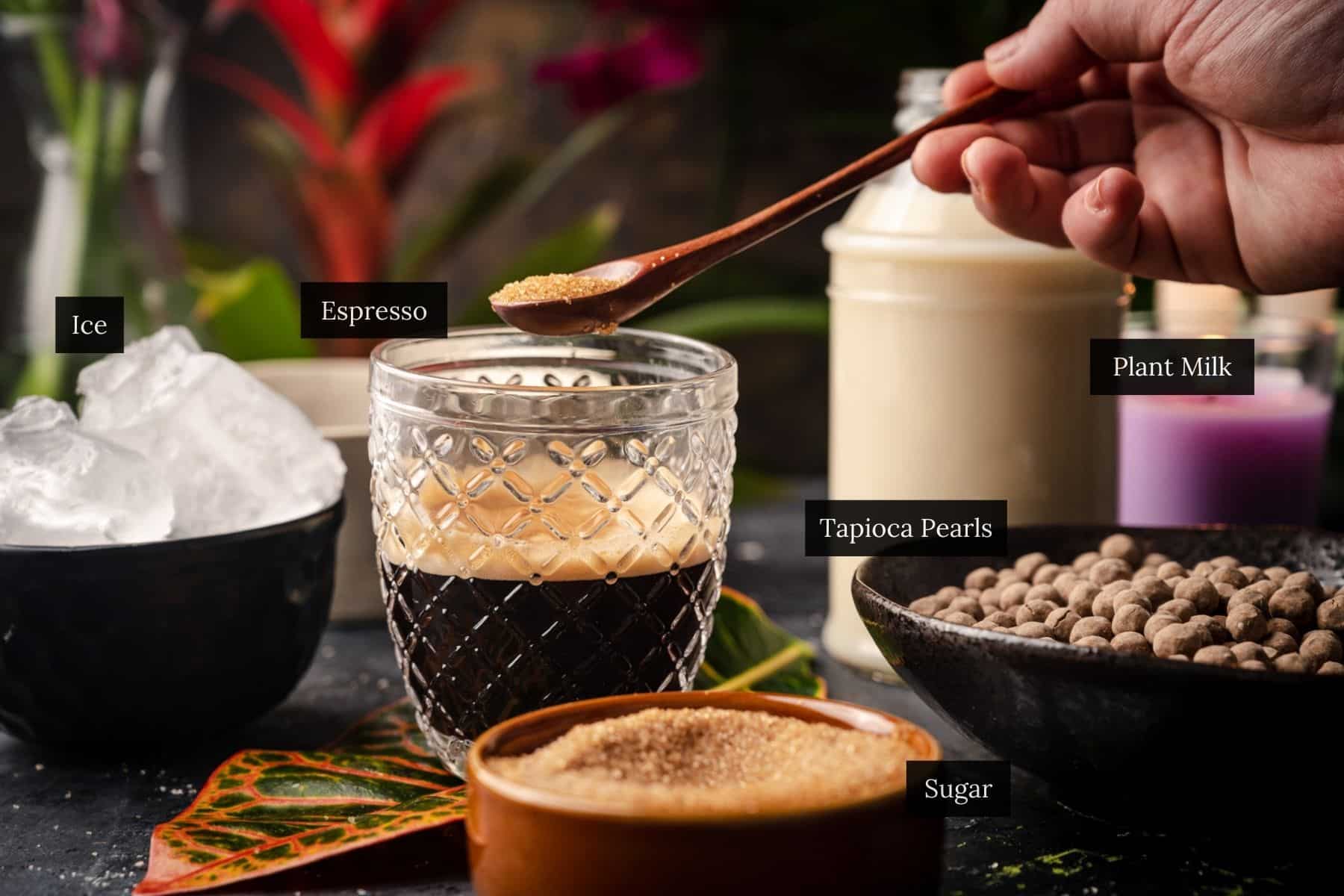 A hand adds sugar to a glass of espresso, crafting a unique coffee boba recipe. Nearby are bowls of ice, tapioca pearls, sugar, and a bottle of plant milk, ready to complete this delightful fusion.