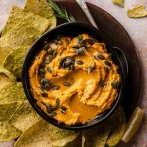 A bowl of creamy pumpkin hummus topped with crunchy pumpkin seeds, accompanied by green tortilla chips on a dark serving board.