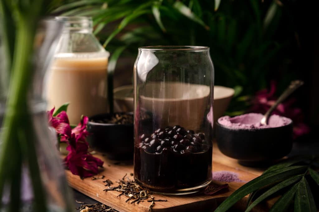 Boba in a clear glass cup with a bowl of freeze dried purple taro behind it.
