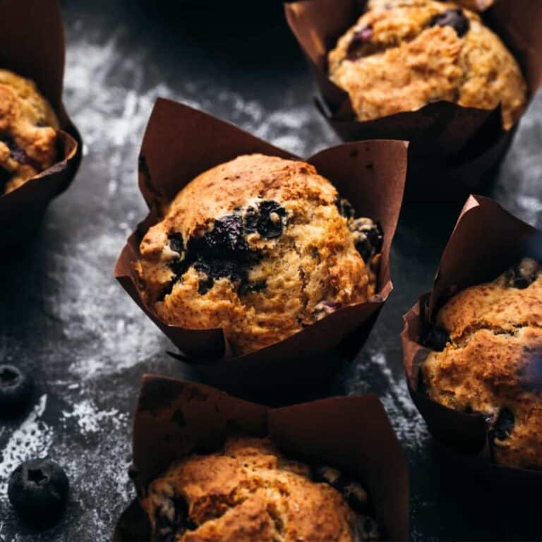 Several freshly baked vegan blueberry muffins in brown paper liners on a dark surface with scattered blueberries.