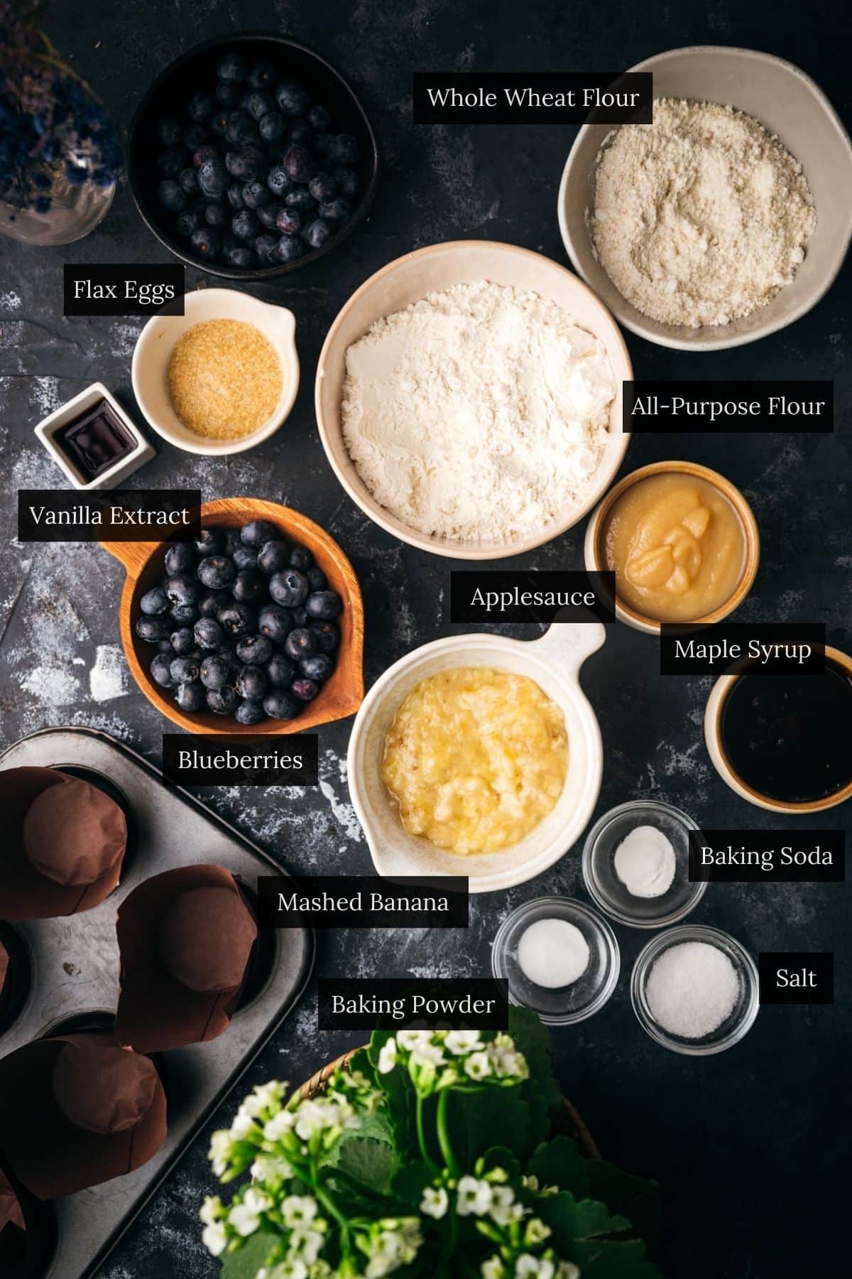 Overhead view of labeled baking ingredients for vegan blueberry muffins: whole wheat flour, all-purpose flour, flax eggs, applesauce, maple syrup, mashed banana, baking soda, baking powder, salt, vanilla extract, blueberries.