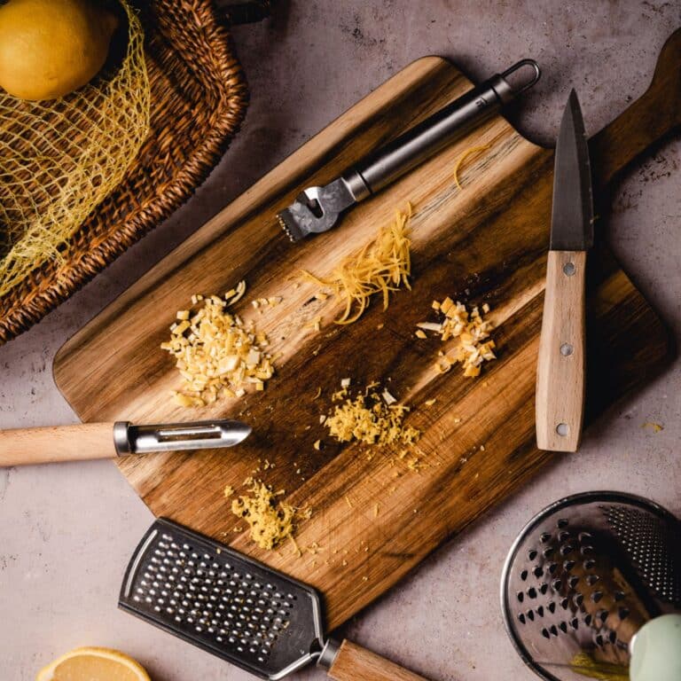 Wooden cutting board with grated pieces of lemon zest, a peeler, knife, grater, and lemon.