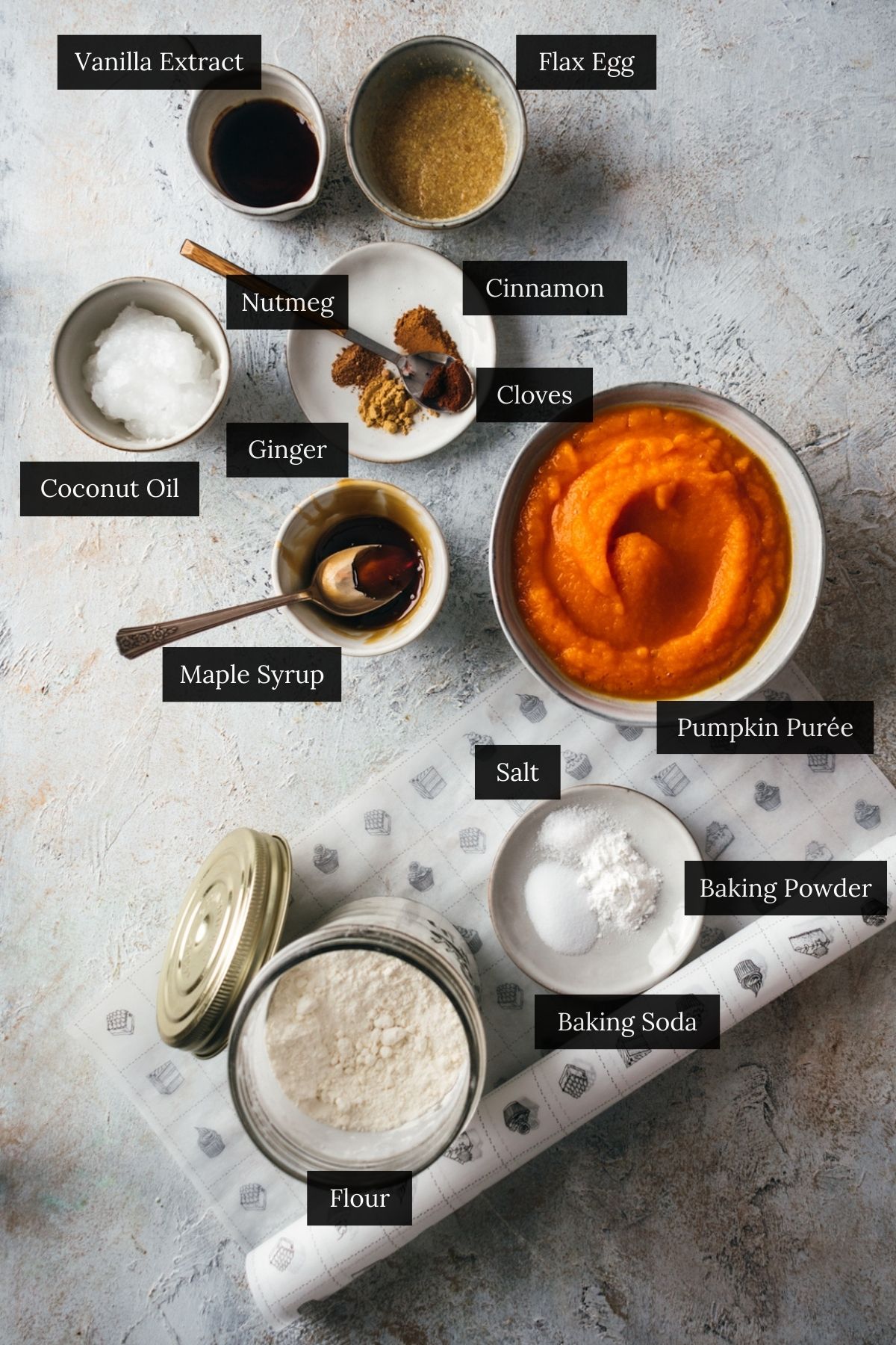 Ingredients for pumpkin scones laid out on a light-colored surface include vanilla extract, flax egg, nutmeg, cinnamon, coconut oil, ginger, cloves, pumpkin purée, maple syrup, salt, baking powder, baking soda and flour.