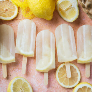 Five lemon popsicles with a lemon slice inside, arranged on a pink surface surrounded by sliced and whole lemons.