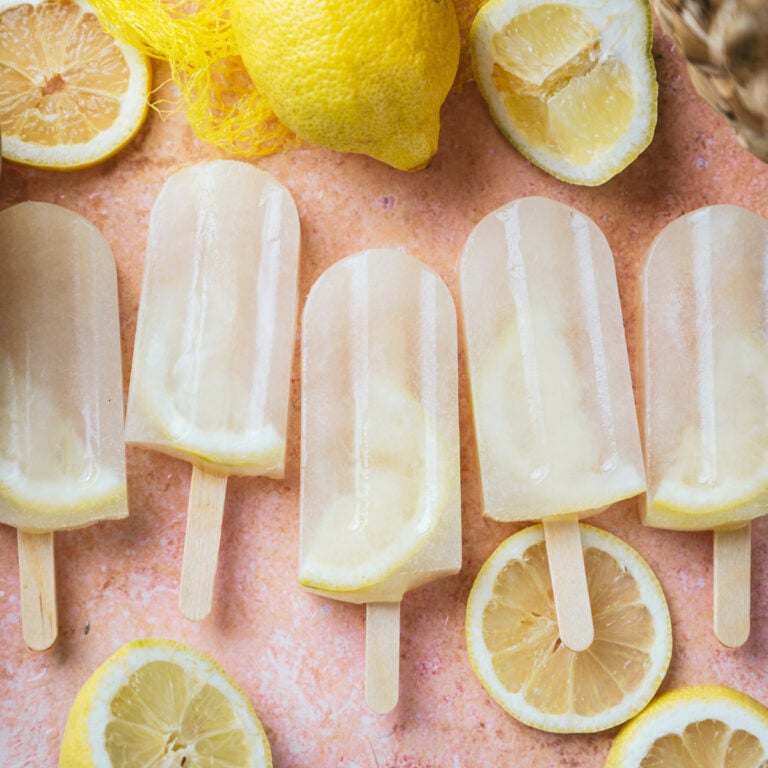 Five lemon popsicles with a lemon slice inside, arranged on a pink surface surrounded by sliced and whole lemons.