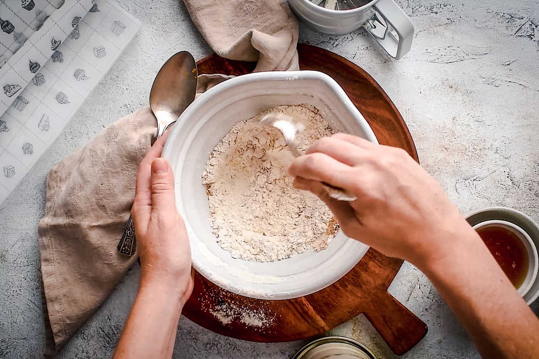 Hands mixing flour in a white bowl with a spoon on a wooden board.
