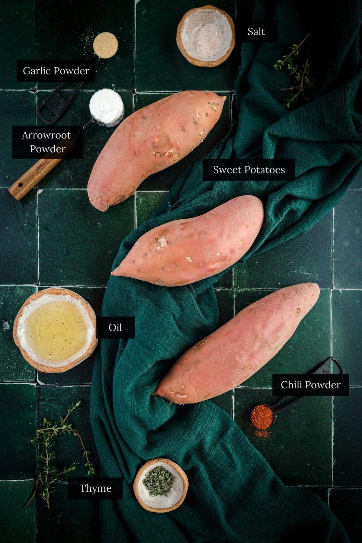 Three sweet potatoes on a green tiled table, surrounded by small bowls labeled garlic powder, arrowroot powder, salt, chili powder, thyme, and oil.