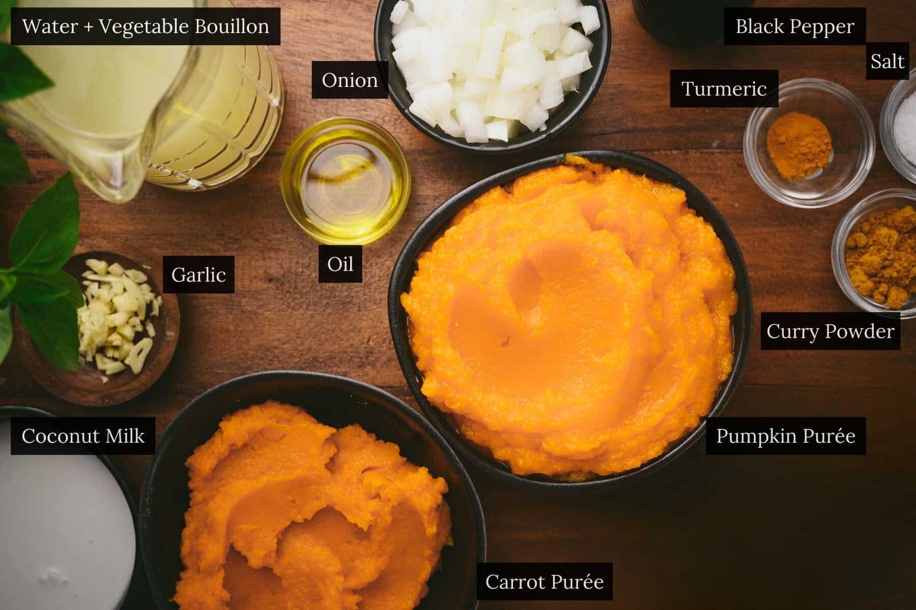Ingredients for a pumpkin curry soup recipe are arranged on a wooden surface, featuring carrot purée, pumpkin purée, onion, garlic, oil, coconut milk, spices, and a jug of vegetable broth.
