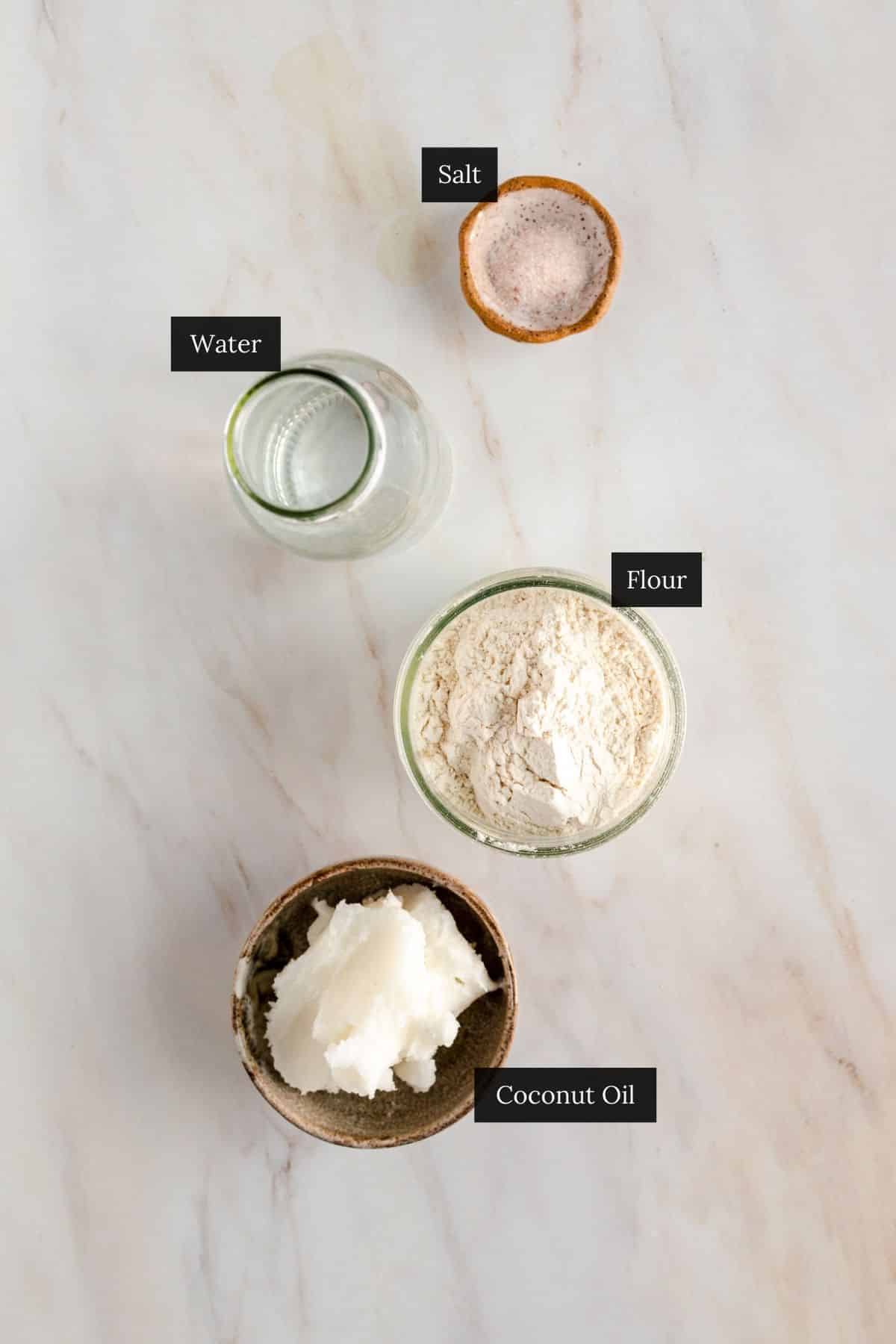 Ingredients on a white surface: a jar of water, a bowl of salt, a bowl of flour, and a bowl of coconut oil, each labeled. Perfect for making a vegan pie crust with coconut oil.