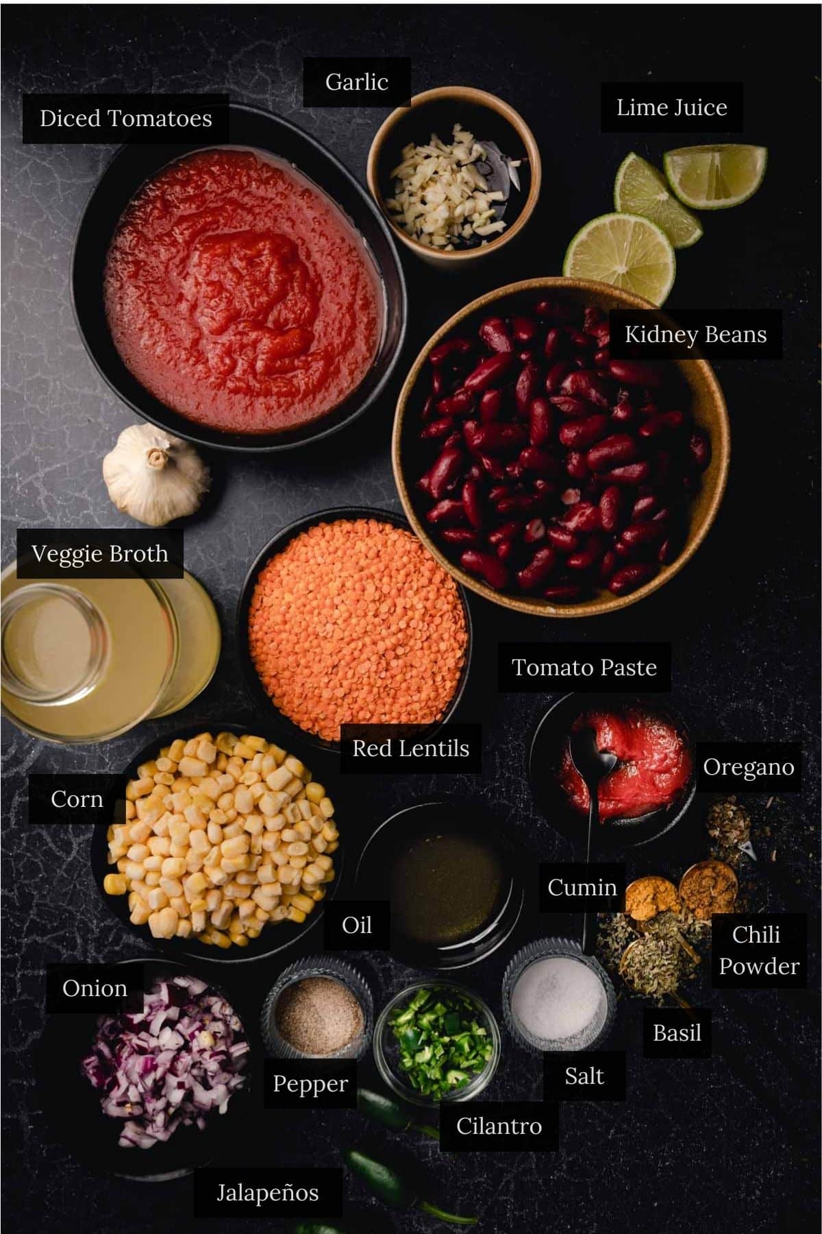 Ingredients for a hearty vegan chili displayed on a black surface: diced tomatoes, garlic, lime juice, kidney beans, veggie broth, red lentils, tomato paste, corn, onion, jalapeños, and various spices.