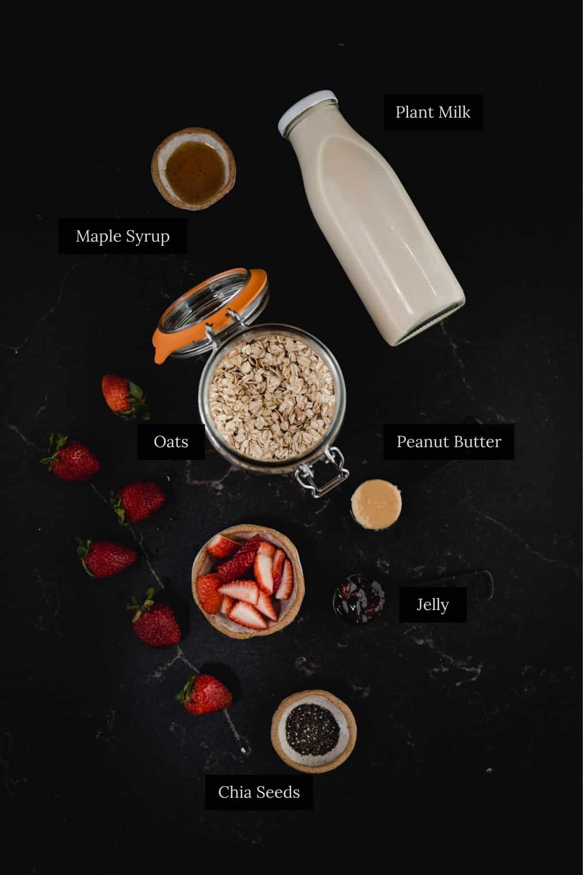 Overhead view of breakfast ingredients: plant milk, oats, chia seeds, strawberries, peanut butter and jelly for delicious overnight oats.