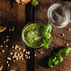 A jar of vibrant vegan pesto sits on a wooden surface, surrounded by scattered pine nuts and fresh basil leaves. A lemon wedge and bread crusts are visible nearby, making this an easy 5-minute recipe for a delightful snack.