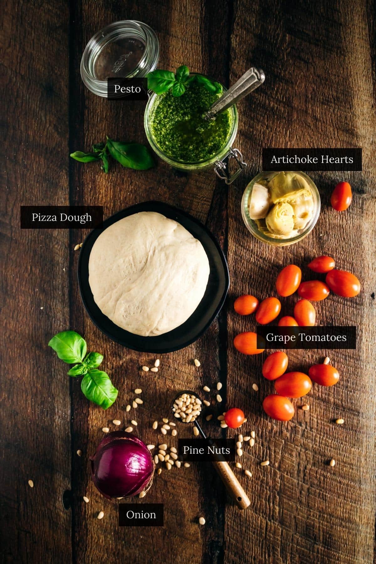 Ingredients on a wooden table: pizza dough, pesto, artichoke hearts, fresh tomatoes, onion, pine nuts, and fresh basil leaves.